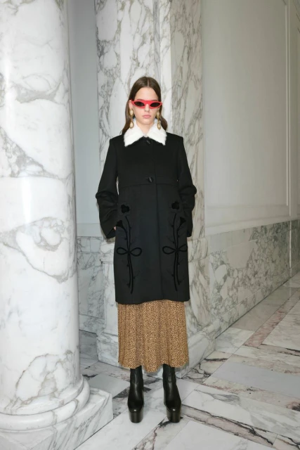 A stylish young woman poses in a black coat and patterned dress against a marble wall backdrop, showcasing modern fashion.