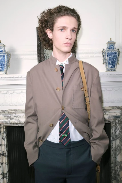 Young adult male with curly hair, posing in a light brown blazer, patterned shirt, and dark pants, with a shoulder bag and a tie.
