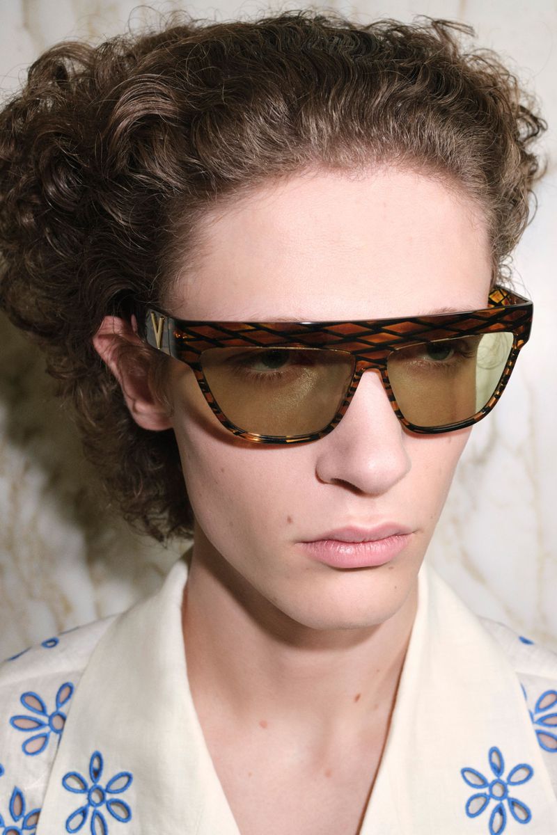Close-up shot of a male model wearing tortoise-shell sunglasses and a patterned collared shirt against a marble background.