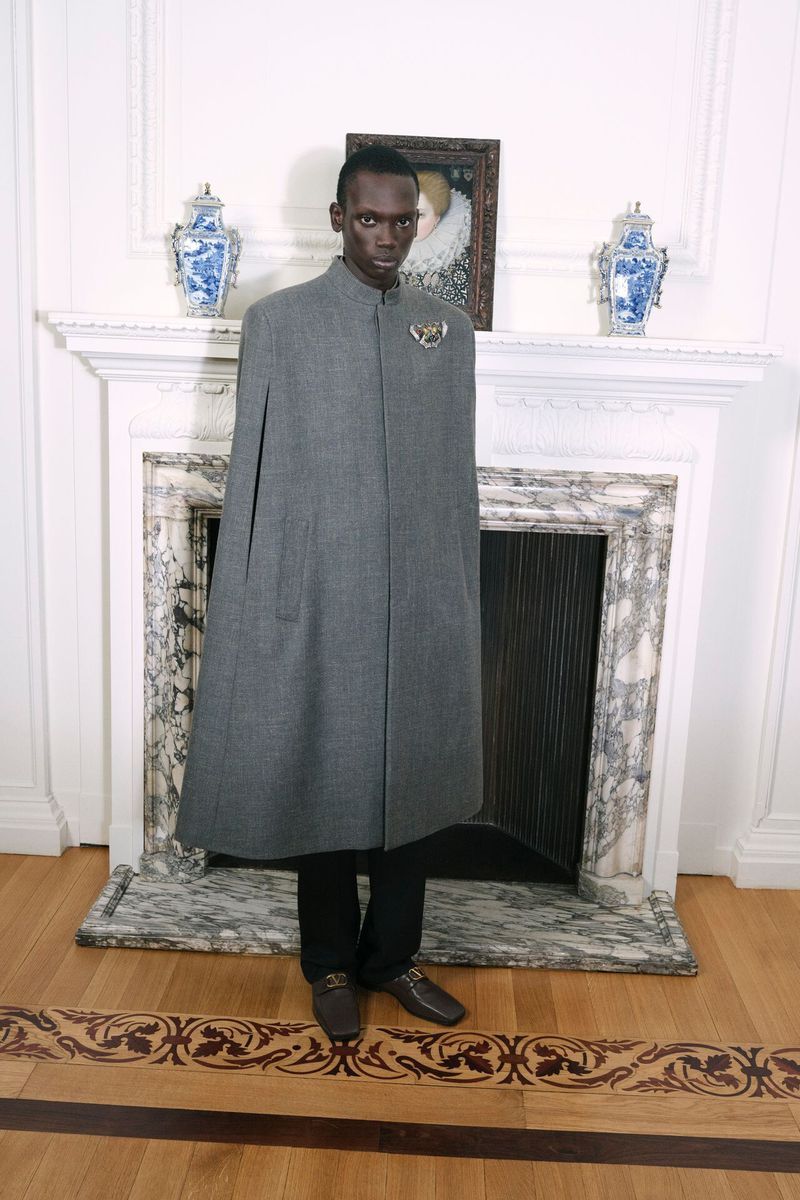 An African-American man stands in a gray cloak in front of a white mantelpiece in an elegant, well-lit room, showcasing a formal style.