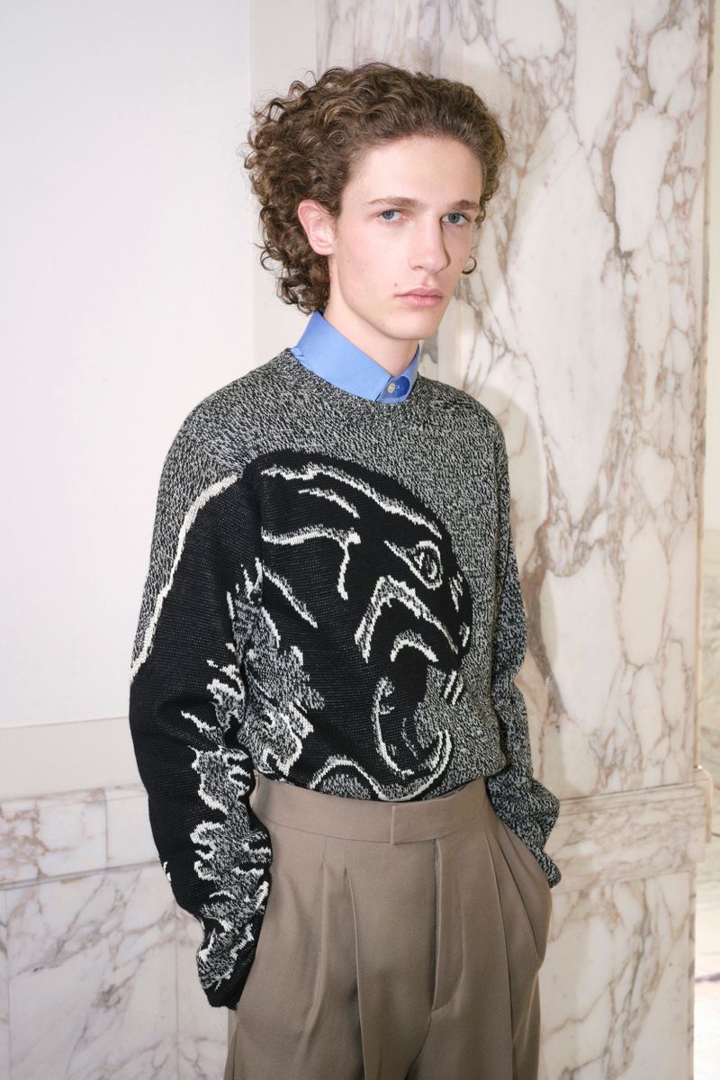 Portrait of a young fair-skinned male model with curly brown hair, wearing a patterned sweater and tan dress pants against a marble background.