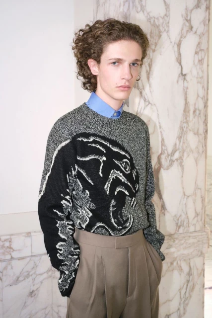 Portrait of a young fair-skinned male model with curly brown hair, wearing a patterned sweater and tan dress pants against a marble background.