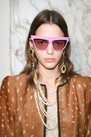Indoor portrait of a young woman wearing pink sunglasses, statement earrings with green gemstones, and multi-strand pearl necklace.