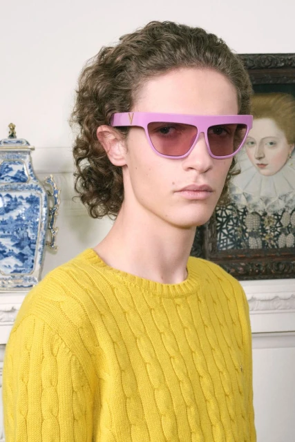 Man with curly brown hair and yellow cable knit sweater poses in front of ornate background wearing wide pink sunglasses.