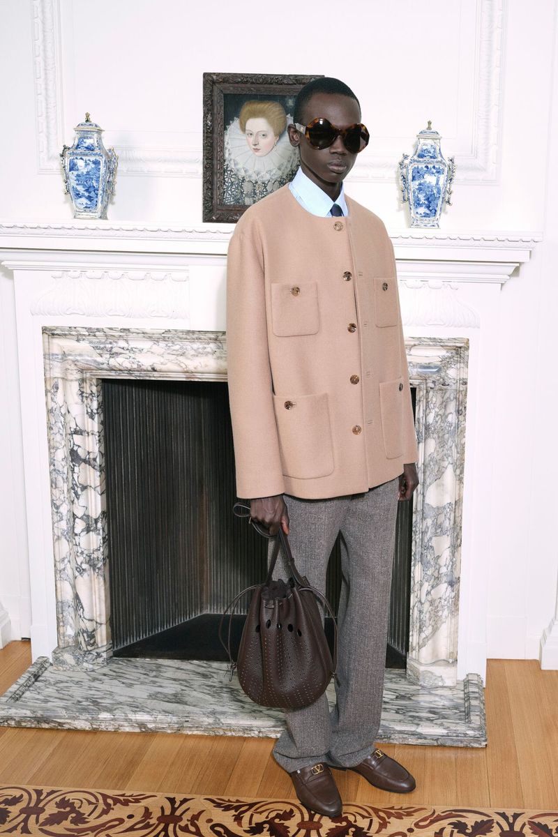 A slender Black man in a tan jacket and grey pants poses indoors, holding a brown leather bucket bag. Portrait art on the wall.
