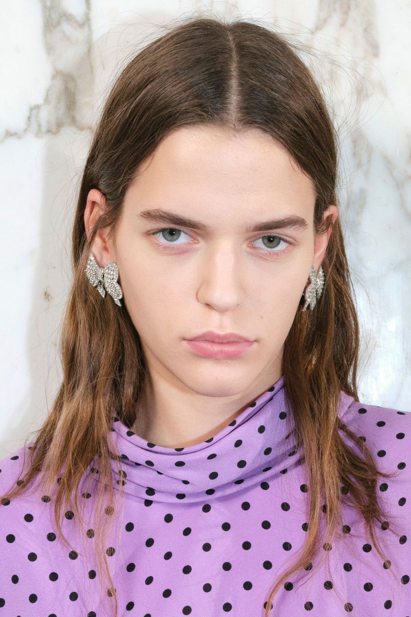 Close-up of a woman with long brown hair wearing crystal earrings and a lavender polka dot top against a marble background.