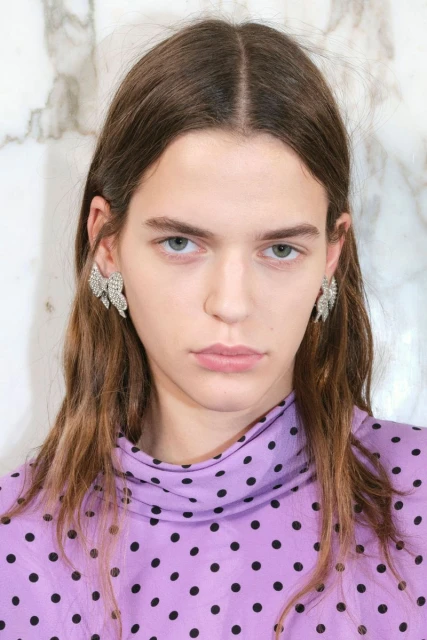 Close-up of a woman with long brown hair wearing crystal earrings and a lavender polka dot top against a marble background.
