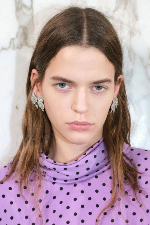 Close-up of a woman with long brown hair wearing crystal earrings and a lavender polka dot top against a marble background.