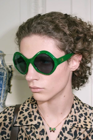 Close-up of a model wearing oversized green textured sunglasses, a leopard-print shirt, and a butterfly necklace.