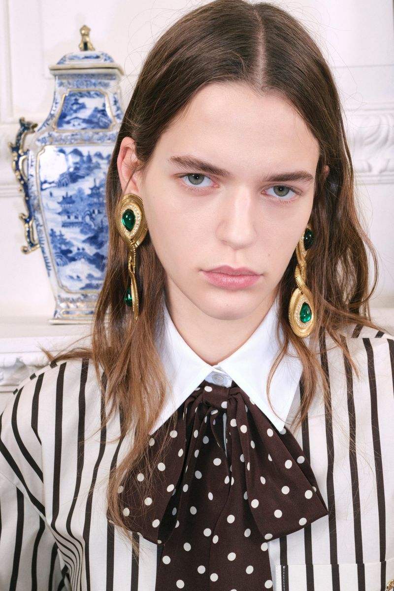 Close-up of a model with long brown hair, wearing a striped blouse with a large polka dot bow and emerald green earrings.