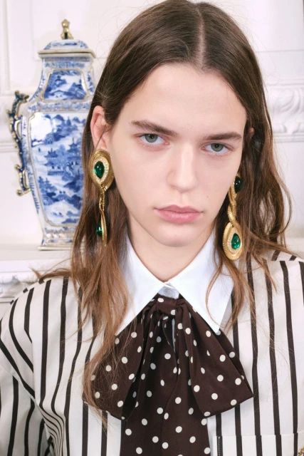 Close-up of a model with long brown hair, wearing a striped blouse with a large polka dot bow and emerald green earrings.
