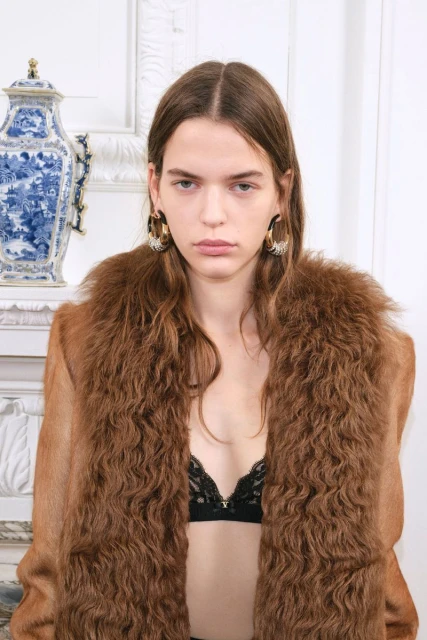 Fashionable brunette woman poses indoors wearing a brown fur coat and black lace lingerie top near a blue and white porcelain vase.