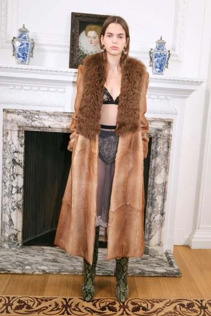 Young woman poses indoors wearing a fur coat, lingerie, and patterned boots against a marble fireplace and an ornate framed portrait.