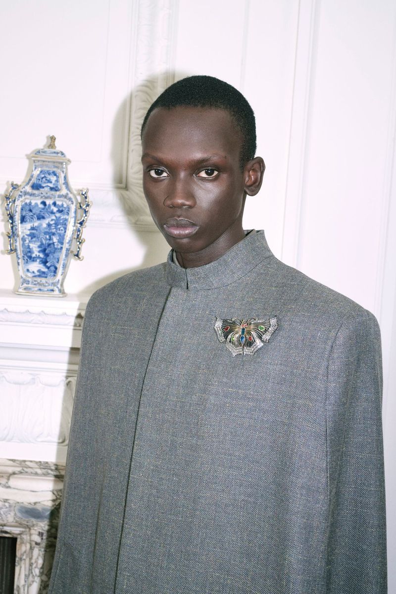 Portrait of a slender black man with serious expression wearing a gray collared cape with ornate brooch in a white paneled room.