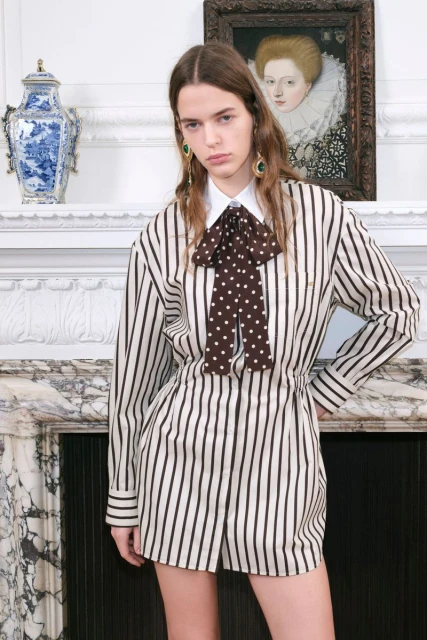 A young woman models a striped romper with a polka-dot bow, posing in front of a white fireplace mantel, framed by an ornate picture and vase.