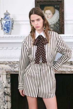 A young woman models a striped romper with a polka-dot bow, posing in front of a white fireplace mantel, framed by an ornate picture and vase.