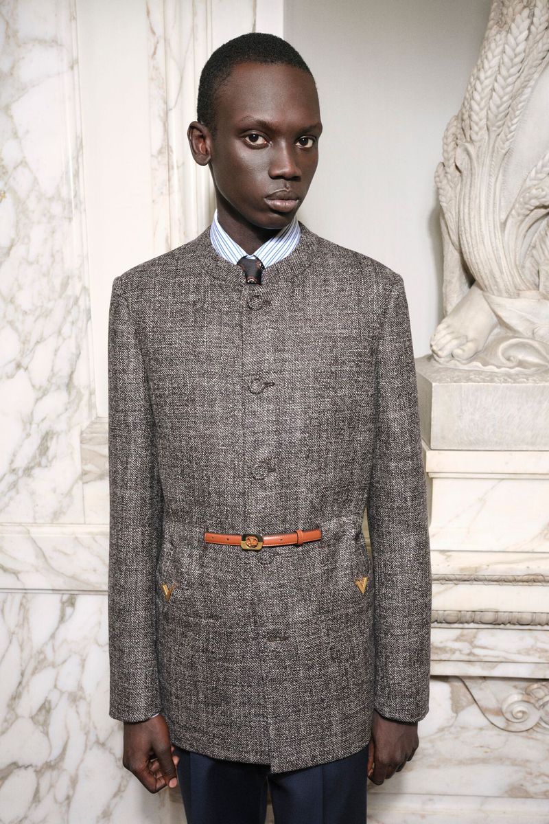 A singular adult male poses in an indoor setting, wearing a grey herringbone jacket with a tan belt, blue dress pants, and a striped shirt.
