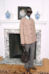 Model wearing a camel jacket and grey trousers standing in front of an ornate white fireplace holding a dark leather bucket bag.