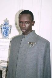 Bukwop Kir wearing a grey high-neck coat with an ornate butterfly brooch against a white wall with a blue porcelain vase.