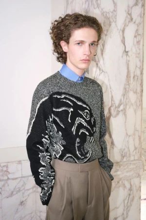 Model with curly hair in a black and grey knit panther sweater with a blue collared shirt and beige pleated trousers by a marble wall.