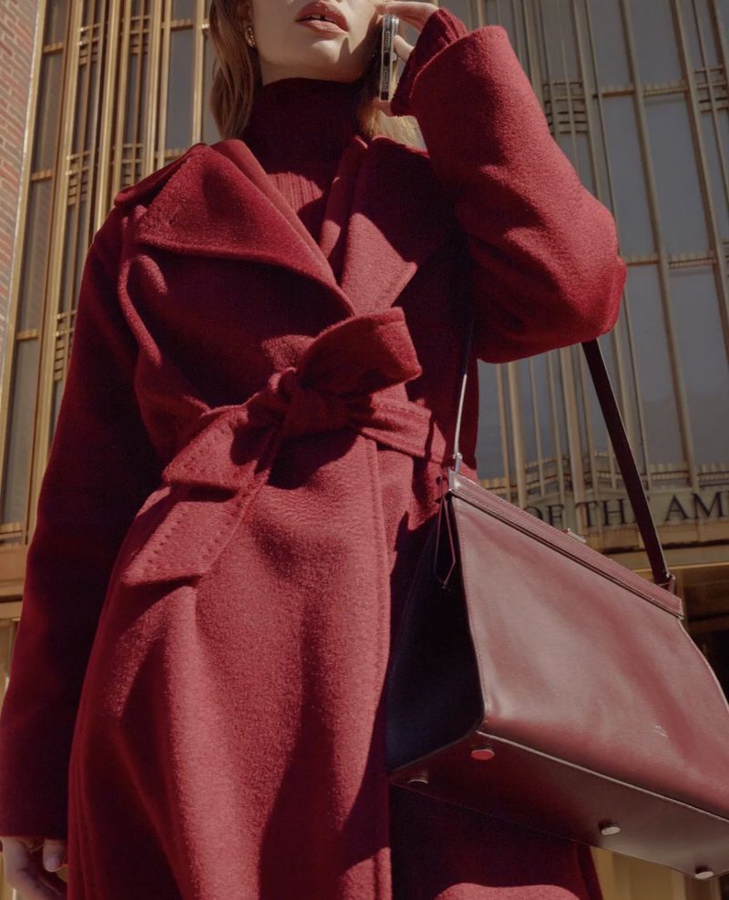 A woman in a red coat talks on her cellphone in front of a building with vertical metal bars and a burgundy leather purse.