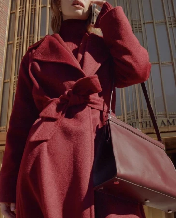 A woman in a red coat talks on her cellphone in front of a building with vertical metal bars and a burgundy leather purse.