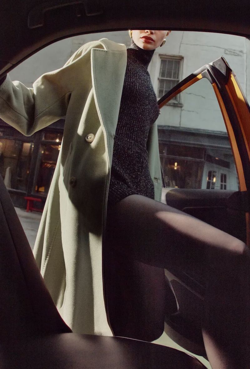Woman in black bodysuit and coat exits car in city, capturing fashion and urban lifestyle with street view in soft, natural light.