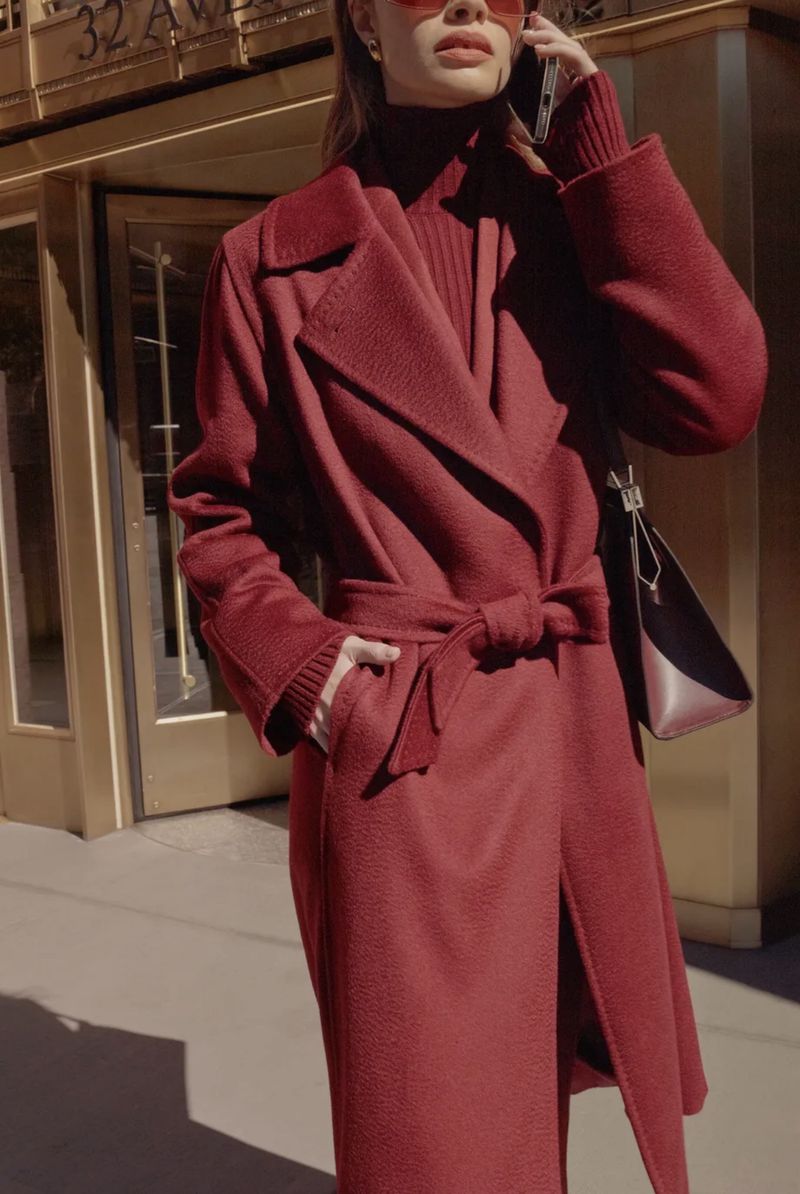 Woman in a red coat speaks on a cell phone outside a building on a sunny day, wearing red sunglasses and a matching sweater and purse.