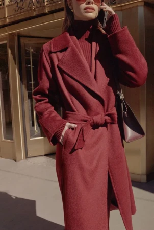 Woman in a red coat speaks on a cell phone outside a building on a sunny day, wearing red sunglasses and a matching sweater and purse.
