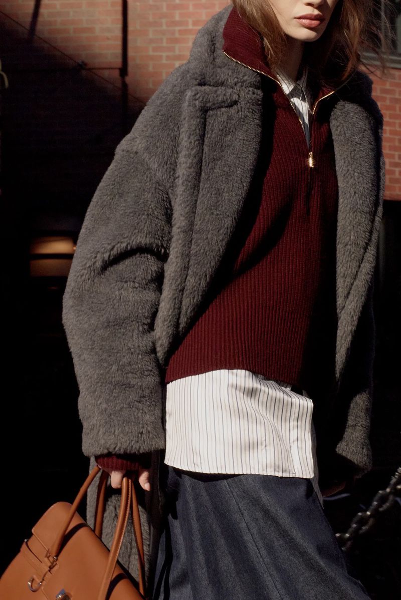 Woman wearing a gray overcoat and holding a tan leather purse stands against a brick wall in an outdoor urban setting.