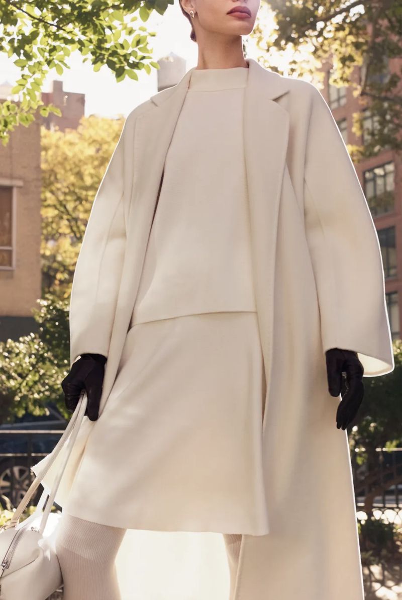 A woman in a cream-colored coat and dress stands on a city street. She is holding a purse and wearing gloves on a sunny day.
