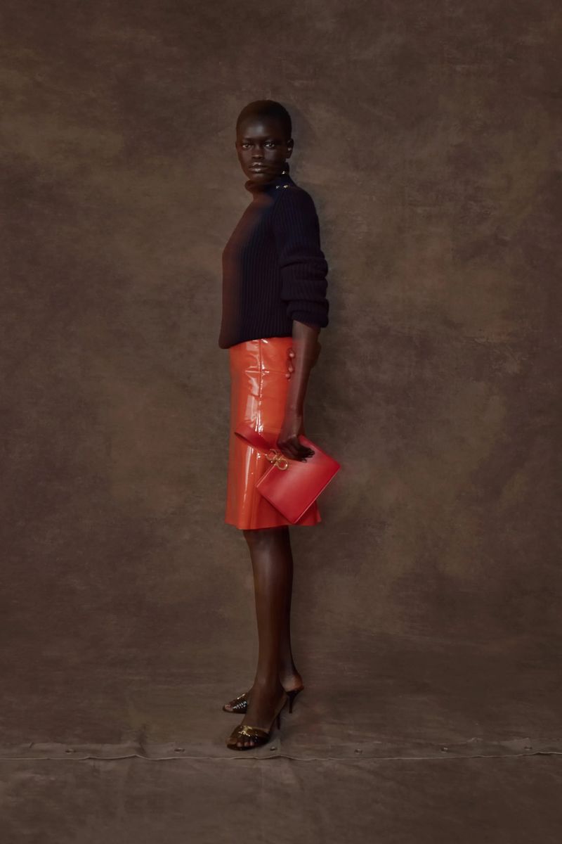 Studio shot of a Black woman in a black turtleneck, orange skirt, and heels holding a red clutch against a brown textured background.