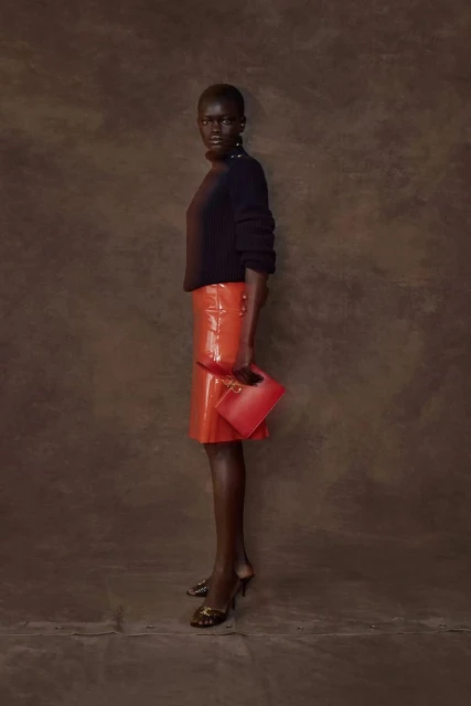 Studio shot of a Black woman in a black turtleneck, orange skirt, and heels holding a red clutch against a brown textured background.