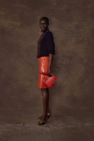 Studio shot of a Black woman in a black turtleneck, orange skirt, and heels holding a red clutch against a brown textured background.