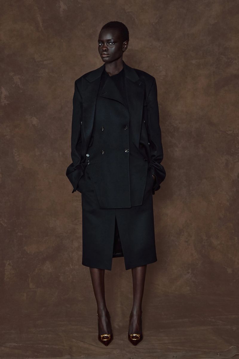 Full-length studio shot of a slender adult woman in a black double-breasted coat, skirt, and heels against a mottled brown backdrop.