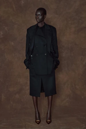 Full-length studio shot of a slender adult woman in a black double-breasted coat, skirt, and heels against a mottled brown backdrop.