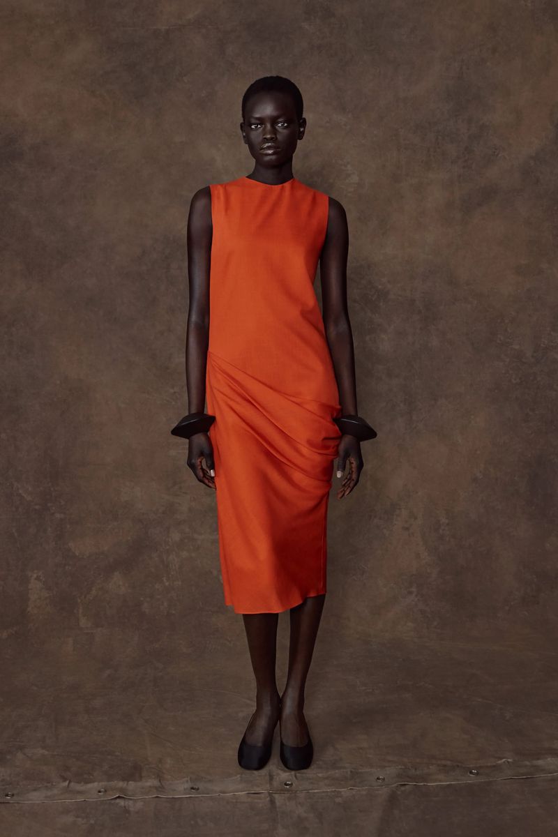Full-length studio shot of a black woman in a sleeveless orange dress, wearing chunky black bracelets, against a brown textured backdrop.