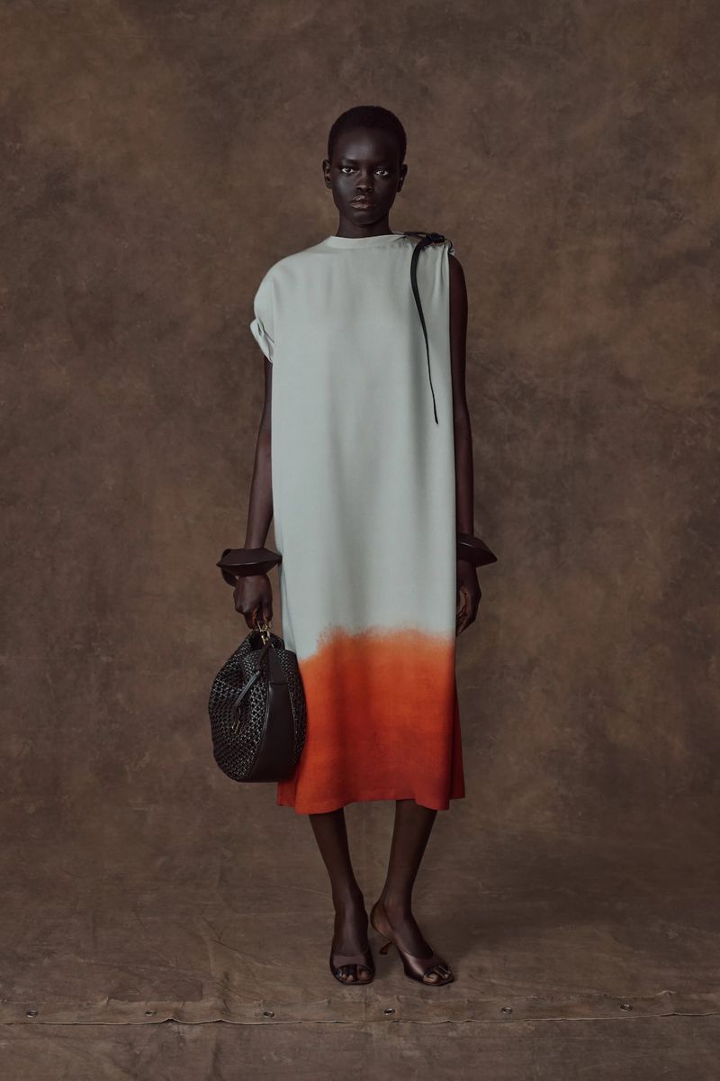 Full-length studio shot of a slender African woman in a neutral dress with a black purse, wearing wooden bracelets against brown backdrop.