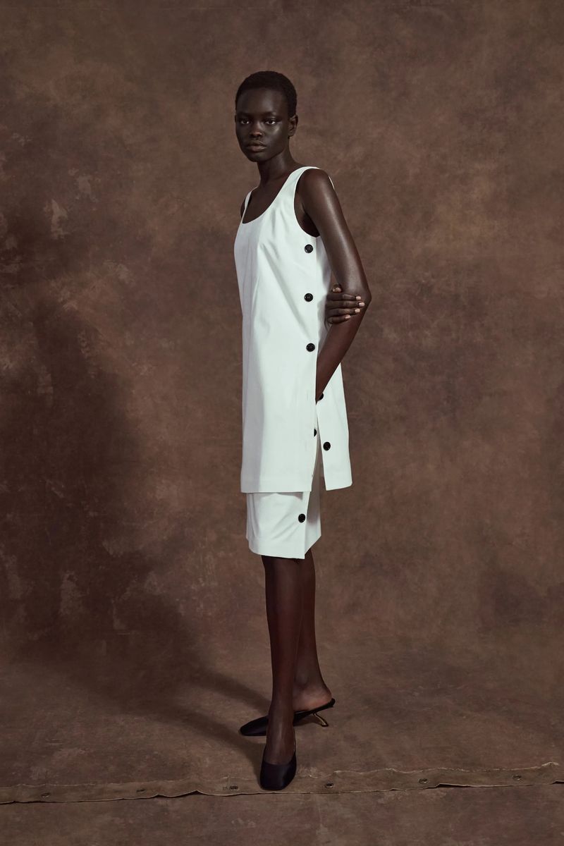 Dark-skinned model posing in a white sleeveless dress with black buttons against a mottled brown backdrop, fashion photography.