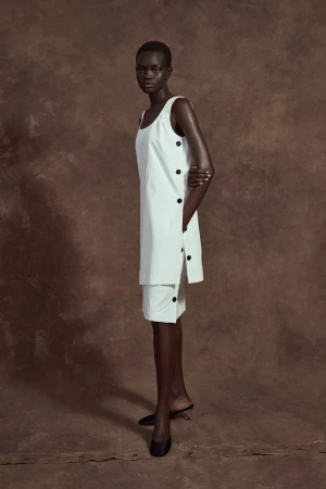 Dark-skinned model posing in a white sleeveless dress with black buttons against a mottled brown backdrop, fashion photography.
