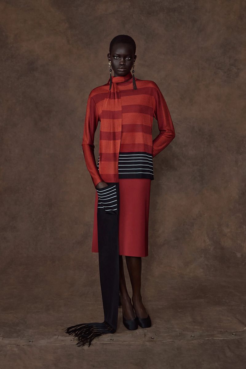 Model Athiec Geng standing in a red and orange striped knit top, matching skirt, and floor-length black scarf against a brown backdrop.