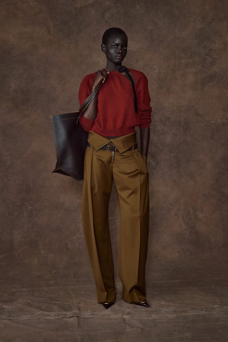 A dark-skinned fashion model poses in a rust-colored sweater and olive trousers with a tote bag against a textured brown backdrop.