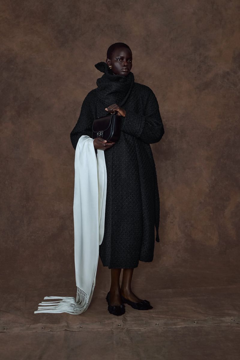 Elegant dark-skinned woman in oversized black coat with white scarf and black purse poses against a textured brown background.