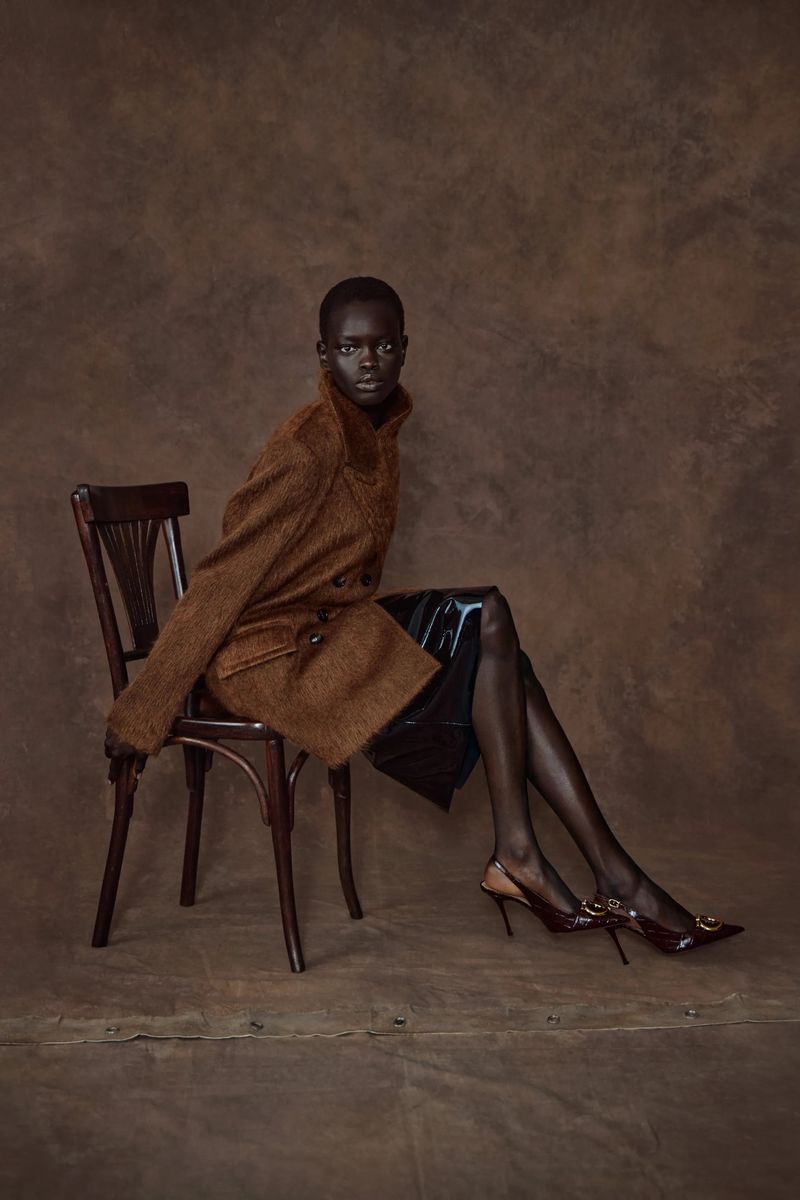 Elegant woman poses in a brown coat and black skirt while seated on a wood chair against a mottled brown backdrop in a studio shot.