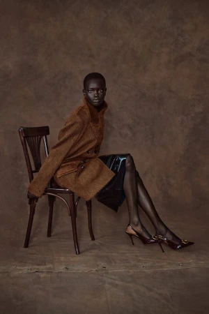 Elegant woman poses in a brown coat and black skirt while seated on a wood chair against a mottled brown backdrop in a studio shot.