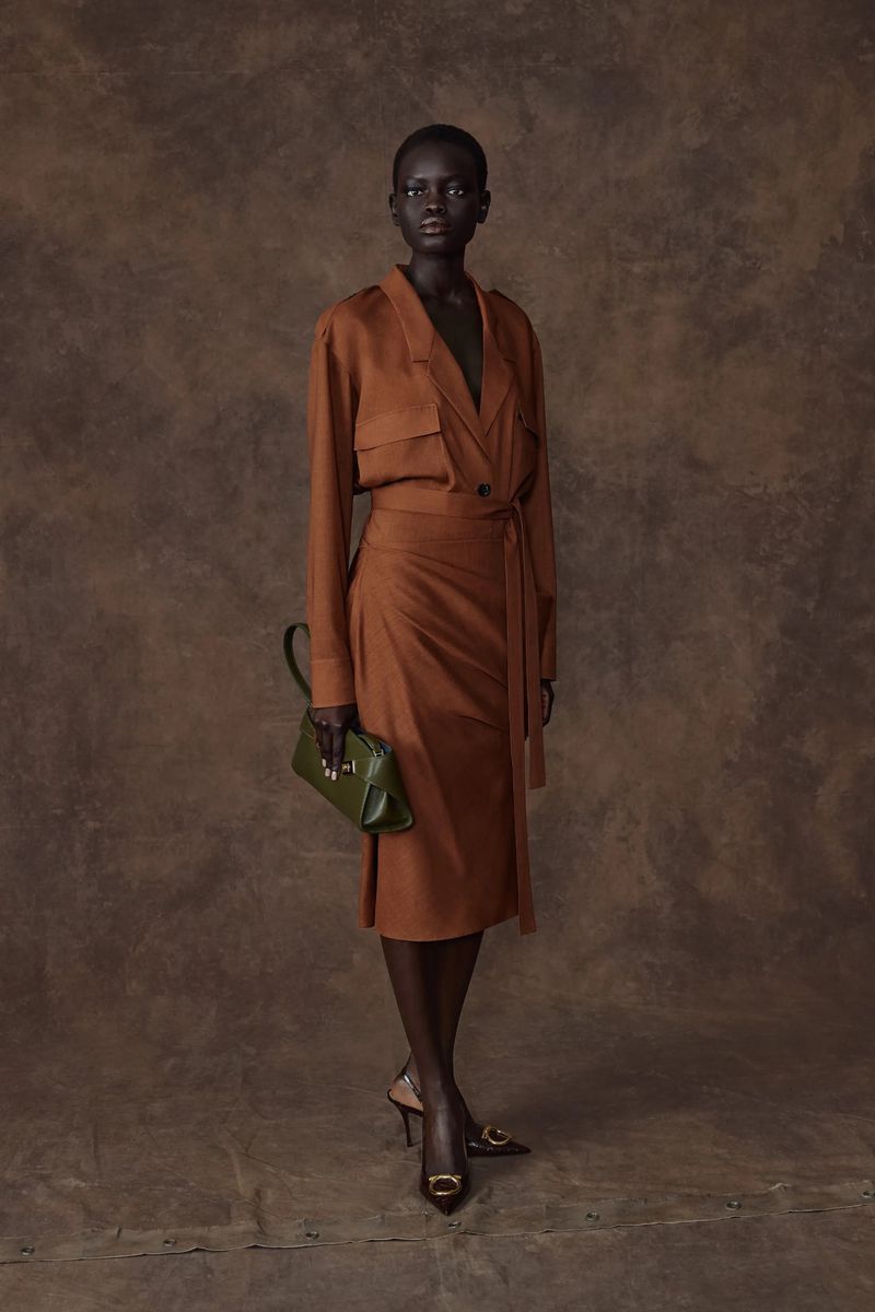 Full-length portrait of a dark-skinned woman in a brown wrap dress and heels, holding a green purse against a mottled brown backdrop.