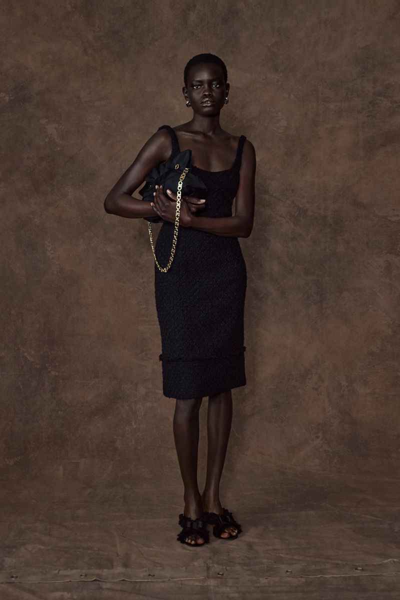 A full studio shot of an African-American woman in a black dress and shoes, holding a black purse with a chain strap against a brown background.