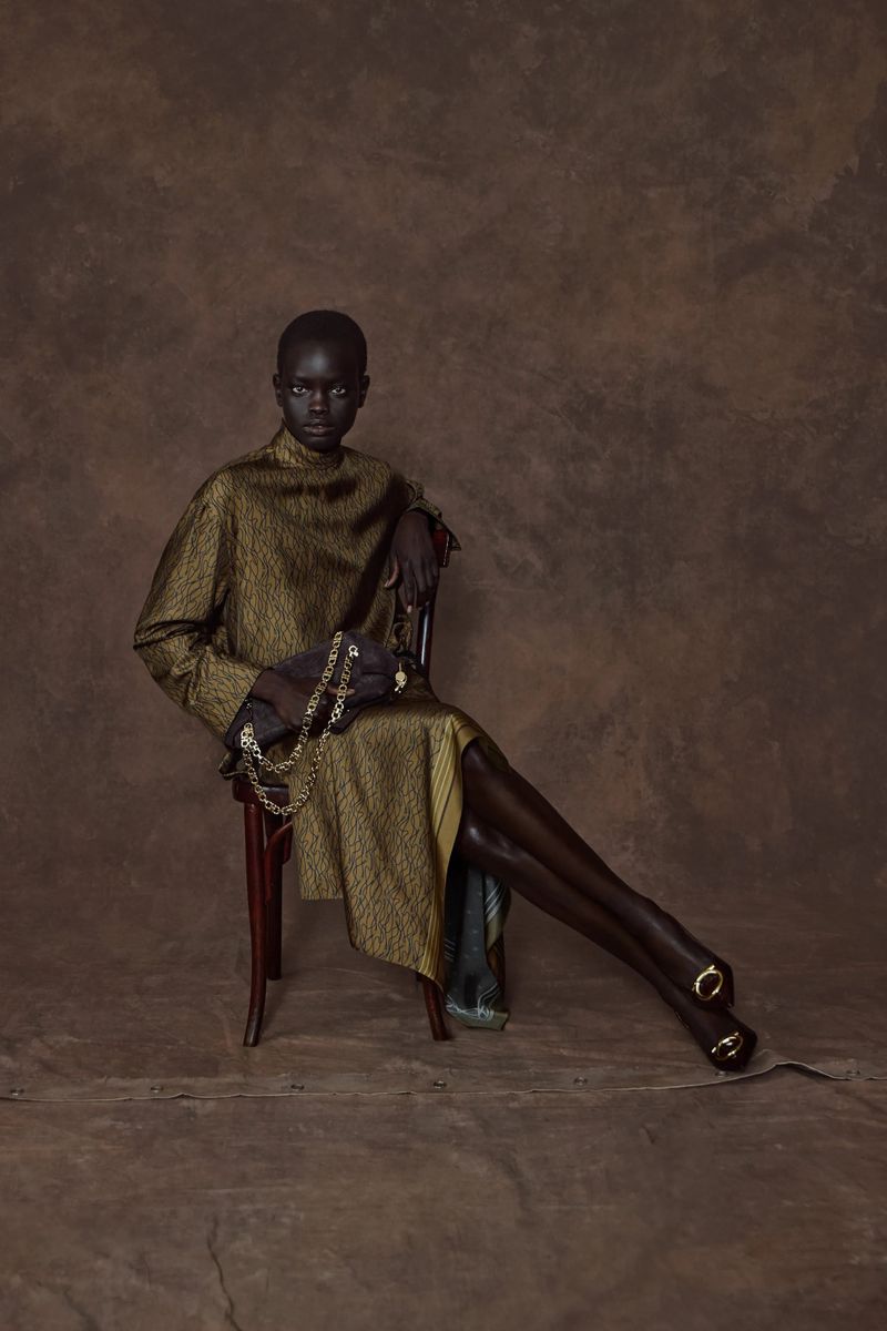 Fashionable black woman in gold dress sitting on chair against brown backdrop holding gold chain. Indoor shot with studio lighting.