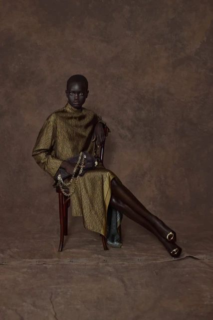 Fashionable black woman in gold dress sitting on chair against brown backdrop holding gold chain. Indoor shot with studio lighting.