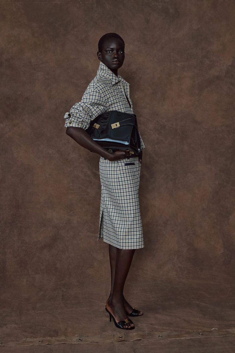 A woman poses in a checkered skirt suit, holding a structured black handbag against a mottled brown background in a fashion editorial shot.